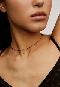 Collar de oro con cuentas negras, que presenta un pequeño colgante de 'B' brillante. Diseño delicado, textura de metal suave y estilo minimalista.