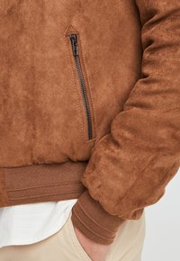 Giubbotto bomber in suede marrone con tasca frontale con zip, polsini a costine e orlo elastico. La texture è morbida con una finitura opaca.