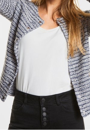 Cardigan en tricot texturé bleu marine et blanc à col rond, fermé par des boutons décoratifs, porté sur un haut blanc et un jean noir taille haute.