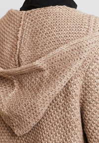 Kardigan z kapturem w jasnobeżowym kolorze wykonany z dzianiny o fakturowanym wzorze. Posiada trójkątny kaptur oraz luźny krój, prezentując miękką przędzę.
