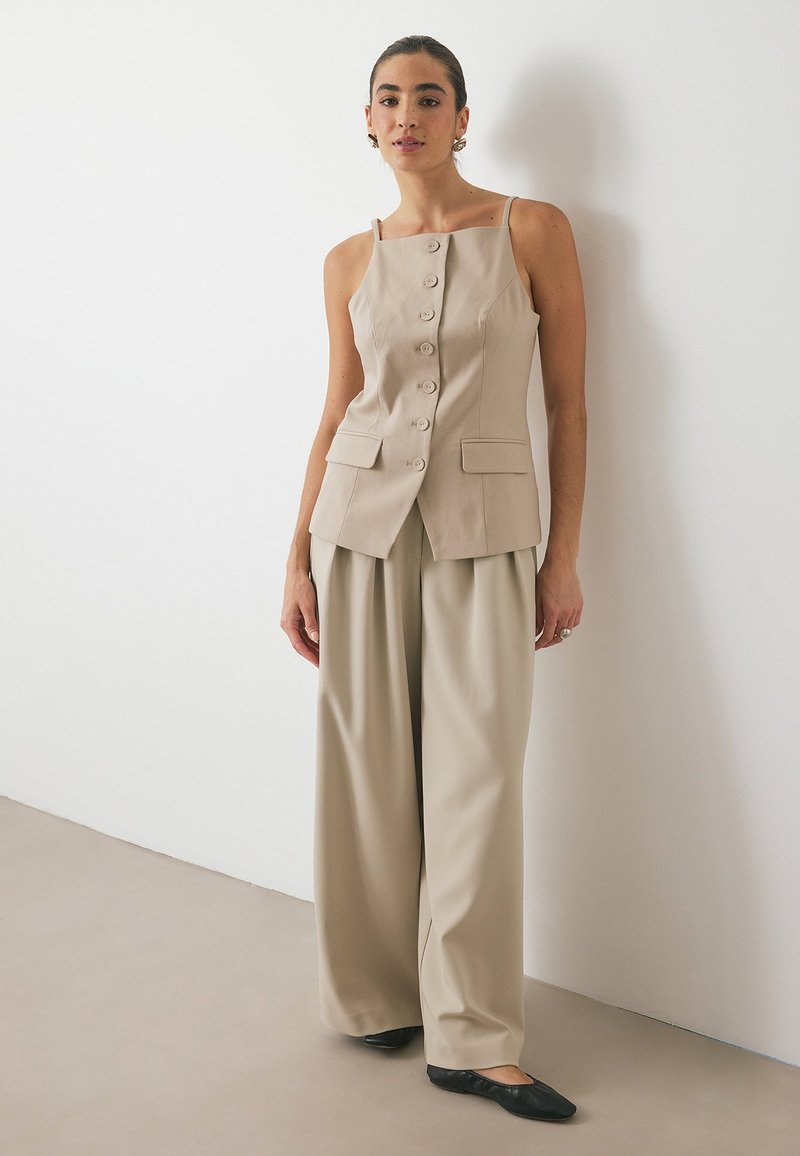 Bež zgornji del brez rokavov z gumbom na sprednji strani in žepi, v kombinaciji z bež širokimi hlačami in črnimi balerinkami. Preprost in prilagojen dizajn.