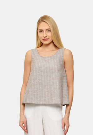 Donna bionda con indosso un top beige a righe senza maniche e pantaloni bianchi larghi, in piedi davanti a uno sfondo chiaro e semplice.
