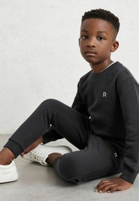 Črn bombažna sweatshirt in trenirke z belim logotipom na prsih in hlačah. Set ima sproščen kroj in rebraste manšete. Obute so bele superge.