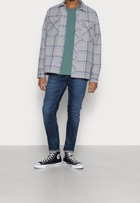 Uomo che indossa una camicia verde, una giacca a quadri grigi, jeans blu scuri e sneaker alte nere, in piedi contro un muro bianco.