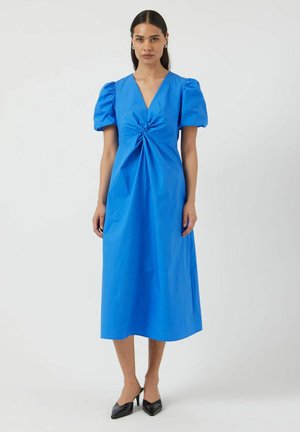 Kvinne iført en knallblå midikjole med puffede korte ermer, V-hals, rynket detalj foran, og svarte spisse hæler.