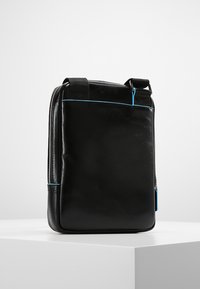 Borsa in pelle nera con un design rettangolare elegante. Caratterizzata da una cerniera blu e una tracolla nera regolabile. Texture liscia.