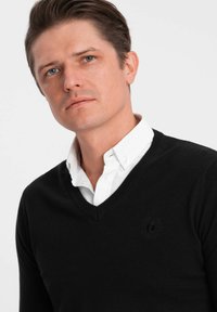 Schwarzer V-Ausschnitt-Pullover aus Strickmaterial, getragen über einem weißen Hemd mit Kragen. Verfügt über ein dezentes gesticktes Logo auf der linken Brust.