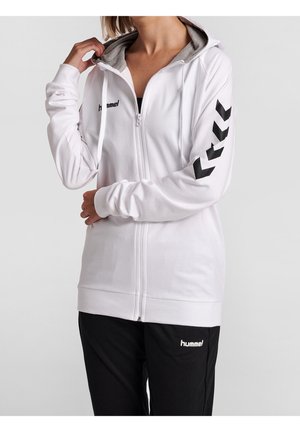 Persoon draagt een witte Hummel-hoodie met rits en zwarte logo's en zwarte broek, hand tilt de capuchon op, bovenlichaam en bovenbenen zichtbaar.