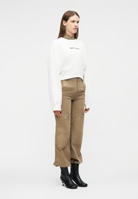 Mujer con sudadera corta blanca, pantalones beige de talle alto con bolsillos delanteros con cremallera, y botas negras de tobillo, de pie contra un fondo liso.