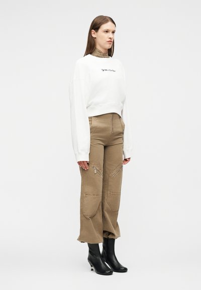 Femme portant un sweat-shirt court blanc, un pantalon beige taille haute avec des poches zippées à l'avant et des bottines noires, debout devant un fond uni.