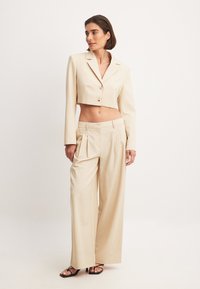 Beige kort jacka med två knappar och långa ärmar, som kombineras med vida byxor med veck och en slät textur. Svarta sandaler bärs.