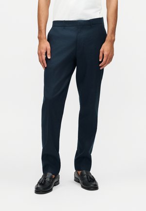 Uomo che indossa pantaloni aderenti blu navy e mocassini neri con nappine, in piedi davanti a uno sfondo bianco uniforme.