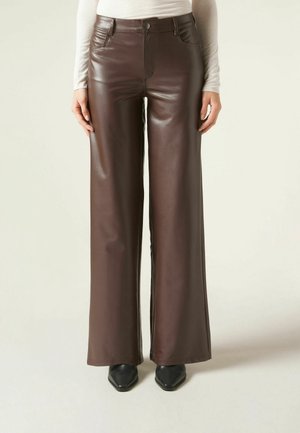 Vista frontale di una persona che indossa pantaloni a gamba larga in ecopelle marrone scuro, scarpe nere con punta affilata e una maglia chiara a maniche lunghe.