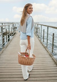 Une femme sourit en marchant sur une jetée en bois au bord de la mer, portant un panier de pique-nique rempli de nourriture et de boissons, vêtue de vêtements légers.