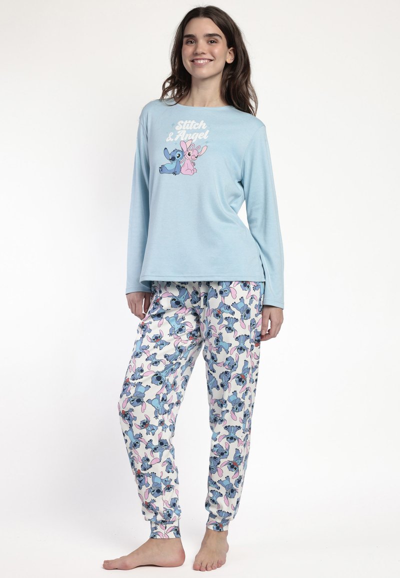 Joven mujer con una camiseta de manga larga azul clara y pantalones de pijama blancos con estampados de personajes de dibujos animados en azul y rosa, de pie descalza.