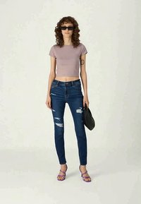 T-shirt cortado roxo, jeans skinny azuis com desgastes, bolsa preta e sandálias roxas. Modelo tem cabelo encaracolado e óculos de sol.