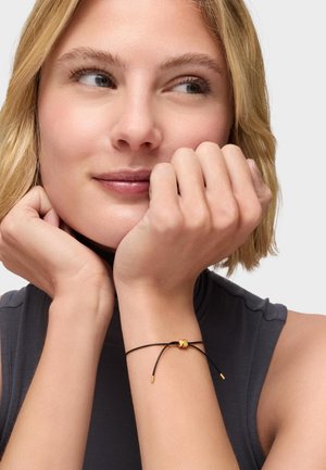 Blondynka z krótkimi włosami w ciemnej, bezrękawnikowej bluzce, opierająca brodę na dłoniach, prezentująca czarną bransoletkę ze złotym ozdobnikiem.