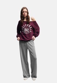 Vinröd sweatshirt med tryckt logotyp, off-shoulder design. Grå bredbenta mjukbyxor. Slät textur, avslappnad passform och ljusfärgad skor.