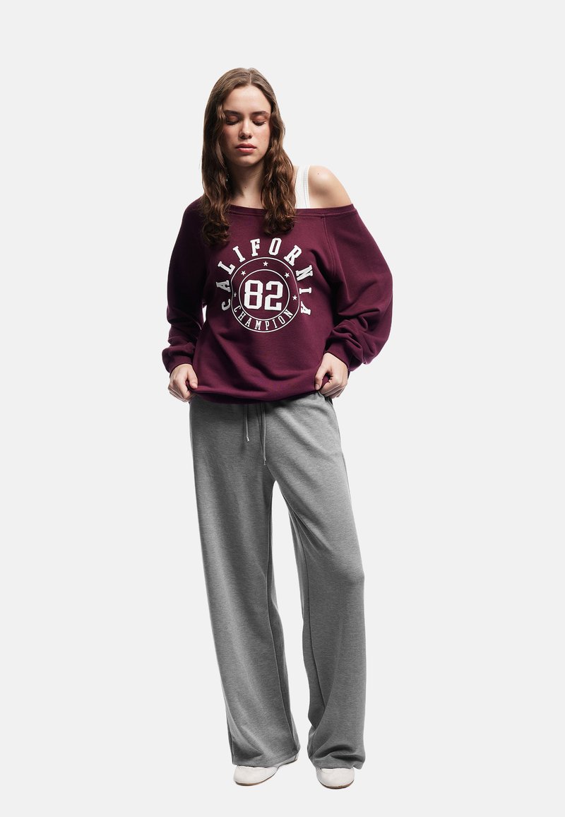 Vinröd sweatshirt med tryckt logotyp, off-shoulder design. Grå bredbenta mjukbyxor. Slät textur, avslappnad passform och ljusfärgad skor.