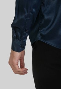 Chemise en satin marine à manches longues, avec des poignets à bouton et une texture lisse. Design ajusté avec un léger éclat visible dans le tissu.