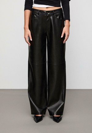 Schwarze weit geschnittene Hose aus Kunstleder mit glatter Textur, vier Taschen und einem Knopfverschluss vorne. Kombiniert mit spitzen schwarzen Absätzen.