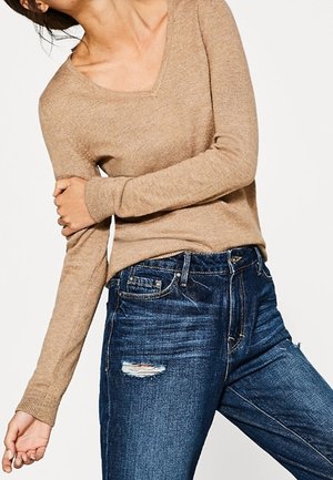 Persona con un suéter beige de cuello en V y jeans azules desgastados, posando con un brazo cruzado y el otro colgando.