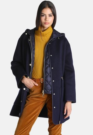Femme portant un manteau à capuche bleu marine, un pull à col roulé côtelé jaune moutarde et un pantalon en velours côtelé marron avec une ceinture en cuir, posant avec une main dans la poche.