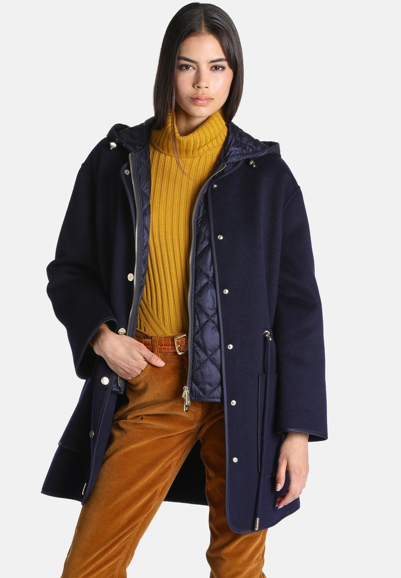 Femme portant un manteau à capuche bleu marine, un pull à col roulé côtelé jaune moutarde et un pantalon en velours côtelé marron avec une ceinture en cuir, posant avec une main dans la poche.