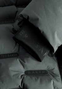 Close-up of a dark padded jacket sleeve with a black elastic cuff embossed with faint text and padded fabric branded with "Ted Baker."