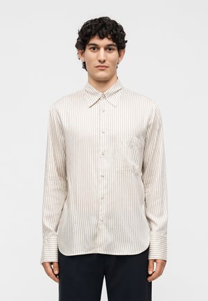 Νέος άνδρας με κοντά σγουρά σκούρα μαλλιά, φορώντας άσπρο πουκάμισο με μακριά μανίκια με λεπτές κάθετες ρίγες και μαύρο παντελόνι.