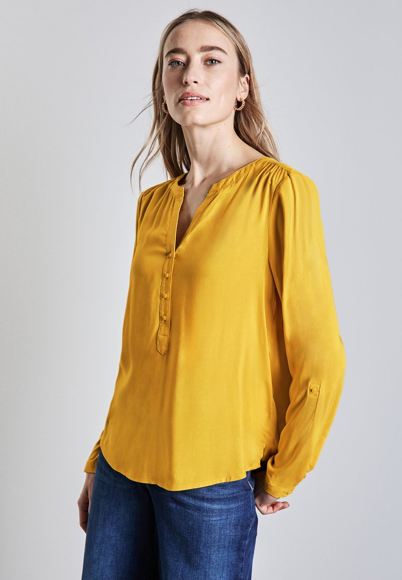 Långärmad gul blus med V-ringning, knappslå och samlade axlar, tillverkad av ett mjukt tyg. Parats med denimjeans.