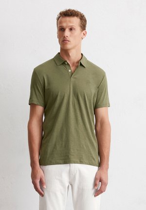Grünes Poloshirt aus weichem Stoff, mit Kragen, zwei Knöpfen und kurzen Ärmeln. Das Modell trägt helle Hosen.