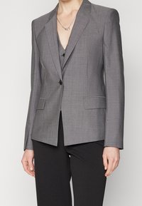 Blazer gris sur mesure avec revers crantés, fermeture à un bouton, et deux poches avant. Confectionné dans un tissu lisse et texturé.