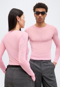 Δύο μοντέλα φοράνε διάφανα ροζ μπλουζάκια με μακριά μανίκια και γκρι καρό παντελόνια, το ένα κοιτάζει μπροστά με γυαλιά ηλίου, το άλλο πλευρικά.