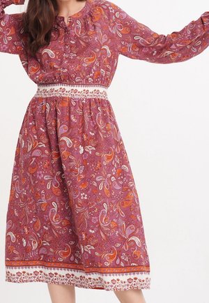 Vrouw draagt een lange jurk met lange mouwen tot op de knie, met een roze en oranje paisley bloemenpatroon en een witte, geborduurde tailleband.