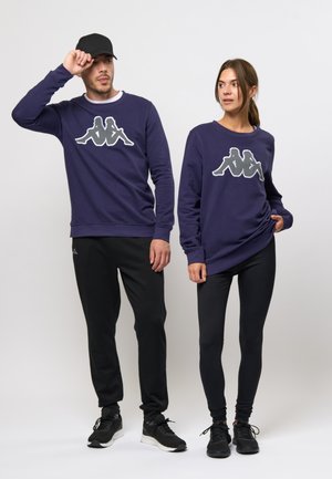 Paarse crewneck sweaters met Kappa-logo in het grijs, gedragen met zwarte leggings en joggers, gecombineerd met zwarte sneakers en een pet.