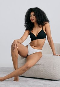 Black wireless bra and white high-cut briefs on a model, featuring smooth fabric and minimalistic design. Model seated on a textured light gray surface.
