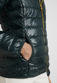 Kort grön puffjacka med kviltat mönster, som har en gul dragkedja och sidofickor. Glansigt, lätt material med mörka detaljer.