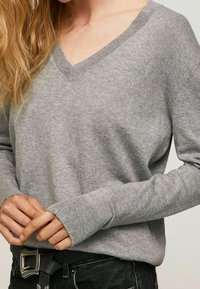 Pull gris à col en V en mati ère tricotée douce. Présente des poignets et un ourlet côtelés, avec une coupe décontractée portée sur un jean noir et une ceinture.