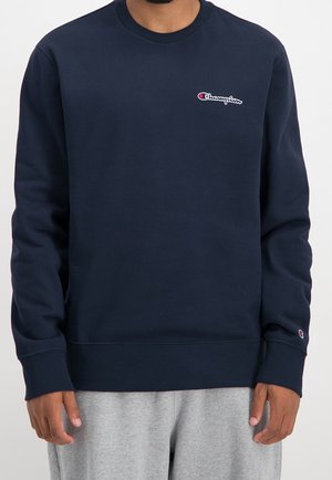 Mann trägt ein marineblaues Champion-Sweatshirt mit Logo auf der linken Brust und am Ärmel, kombiniert mit hellgrauen Jogginghosen.
