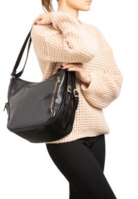 Sac en cuir noir  à l'épaule avec une forme arrondie, doté de ferrures dorées et d'une poche zippée, tenu par une personne portant un pull beige texturé.