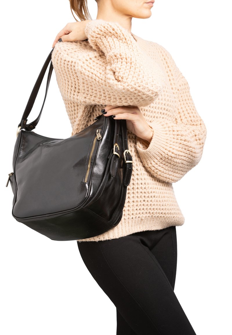 Sac en cuir noir à l'épaule avec une forme arrondie, doté de ferrures dorées et d'une poche zippée, tenu par une personne portant un pull beige texturé.