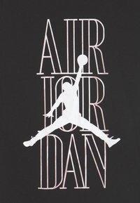 Svart T-shirt med en vit Jumpman-silhuett och fet text "AIR JORDAN" i metallisk roséguld. Enkel grafisk design, bomullsmaterial.