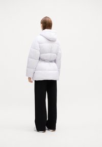 Biela nadýchaná bunda s kapucňou, so priliehavým pásom a logom Calvin Klein na zadnej strane. V kombinácii s čiernymi širokými nohavicami.