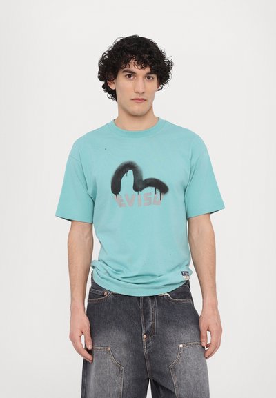 T-shirt in cotone turchese con maniche corte, caratterizzato da un design sprayato di colore nero e il logo "EVISU" in argento metallizzato. Indossato con jeans scuri.