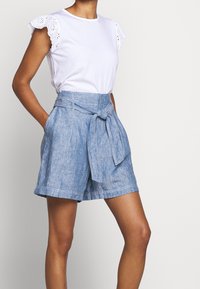 Bílé bavlněné tričko s krajkovými detaily na rukávech, doplněné modrými lněnými šortkami s vysokým pasem, které mají přední zavazování a boční kapsy.