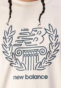 Kremna trenerka z mornarsko grafiko klasičnega stolpa, obdanega z lovorovimi listi in z besedilom "new balance" spodaj.