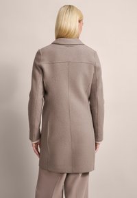 Langer beige Mantel aus strukturiertem Stoff mit Kragen, sichtbaren Nähten und einer leicht ausgestellten Silhouette; kombiniert mit hellen Hosen.