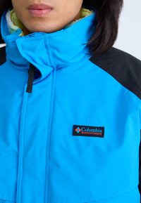 Blaue isolierte Jacke mit hohem Kragen, schwarzen Schulterakzenten und einer weichen, strukturierten Fütterung. Verfügt über ein dezentes Columbia-Logo-Patch.