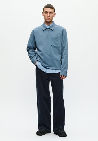 Young man wearing a blue quarter-zip pullover over a striped shirt, dark wide-leg pants, and black shoes, standing with one hand in pocket.